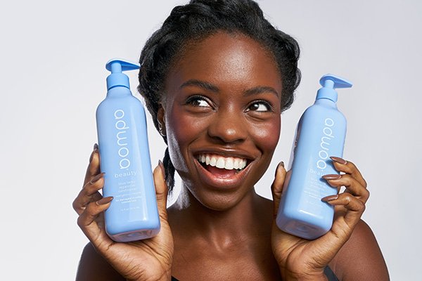 A medium shot of a model holding two bottles of adwoa beauty products to her face and smiling in a studio setting.