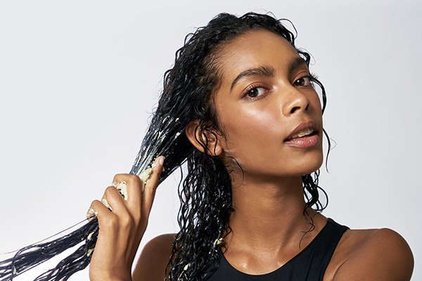 A medium shot of a model adding adwoa beauty product to her long, black, curly hair while looking into the camera. 