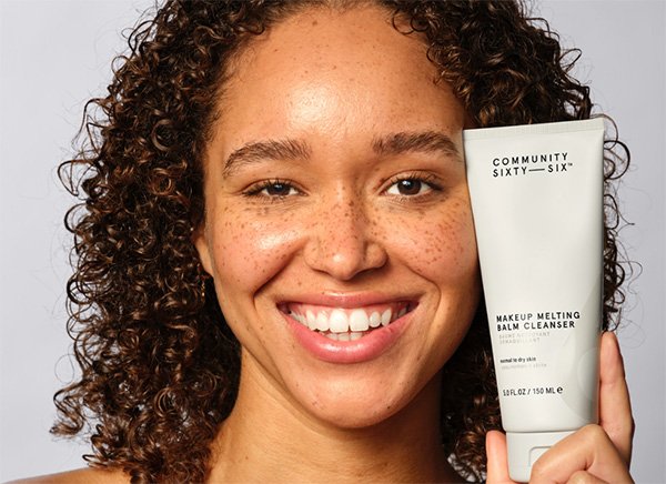 A close up of a woman model with curly hair and freckles looking into the camera and smiling, while holding Community Sixty-Six’s products.