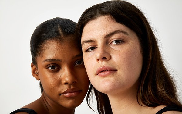 A medium shot of two females models looking into the camera bare faced, in a studio setting on a white background.
