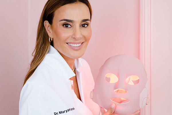 A headshot image of MZ Skin founder Dr. Maryam Zamani in her lab coat holding a LED face mask, in front of a pink wall.