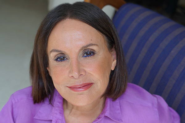 A headshot of BY TERRY's founder Terry de Gunzburg sitting on a stripped chair, wearing a purple shirt looking at the camera. 