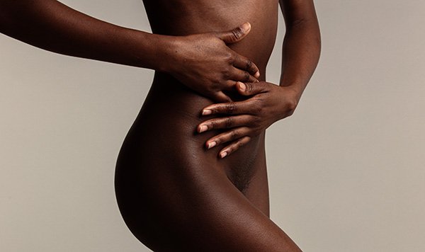 A photo of a slim Black woman in a studio against a grey background, she is naked with both hands placed over her tummy area