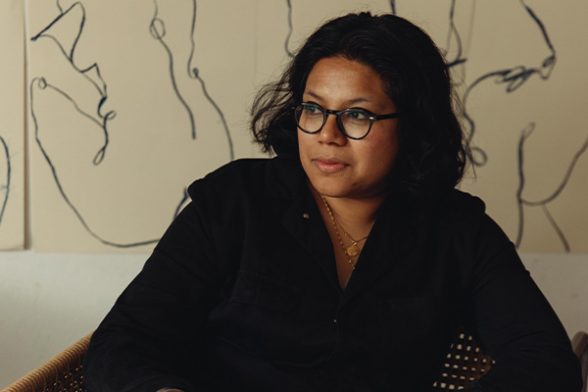 Artist Laxmi Hussain wearing black glasses and a black shirt sitting and looking away from the camera with a lined piece of art behind her on cream coloured paper