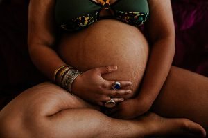 a close up of a pregnant belly being held b a woman wearing a dark green bra and lots of rings in her fingers.