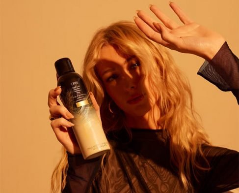 A wide shot image of a blonde-haired model blocking out sunlight while holding hair spray in front of an orange background