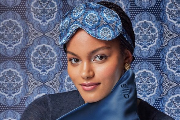 a mid toned model hugging a blue silk pillow and wearing a silk eye mask on her head, shot against a baroque blue print in the background