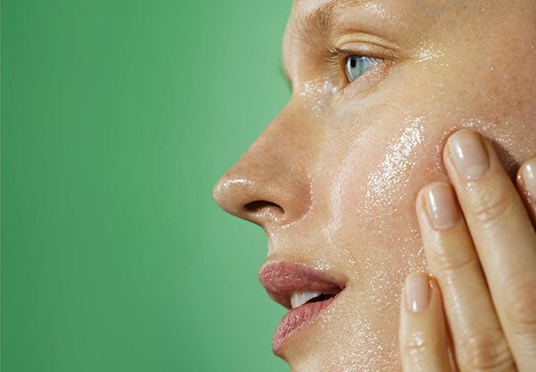 a blonde, fair skinned model applying cleanser to her cheek. the photo is of her left side against a green backdrop