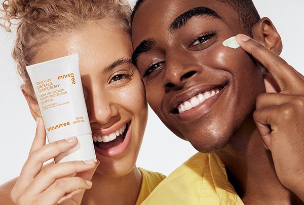 A model smiling holding an innisfree Daily UV Defense Sunscreen, next to another model applying sunscreen on their cheek.