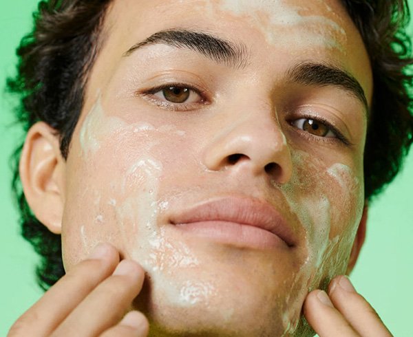 a male olive skinned model with dark curl hair applying a cream cleanser to his face shot against a green background