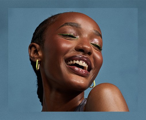 Close up of a dark skinned model against a blue studio background, she wears green eyeliner and lipgloss with her hair in braids