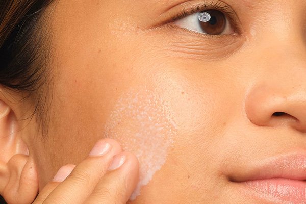 A close up of a model with brown eyes and a tan skin tone looking away and applying a milky skin care formula on her cheek.