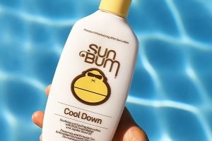 a bottle of sun bum aftersun lotion held above ripples of a swimming pool