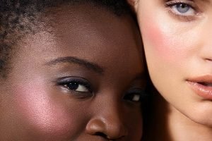 a dark skinned model with short hair and purple shimmery blush sitting next to a fair skinned model wearing the same shimmery blush both looking at the camera