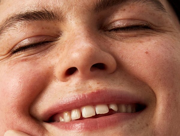 Super close up image of a young woman smiling at the camera with her eyes closed and her top teeth on show in a studio setting