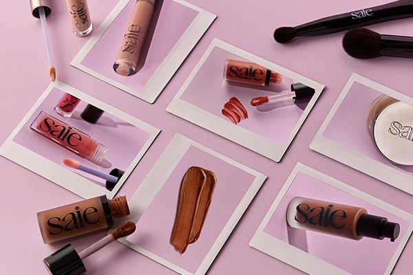Polaroid-like photographs of Saie's signature make up products and beauty brushes on a flat pink surface in a studio setting.