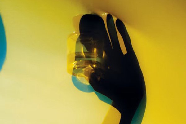A wide shot of a blurred silhouette of a hand holding a bottle of Vyrao fragrance with a yellow background and blue shadows. 