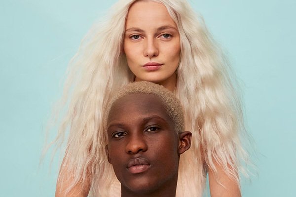 A medium shot of a female and male models with dyed blonde and silver hair in front of a light blue background in a studio setting.