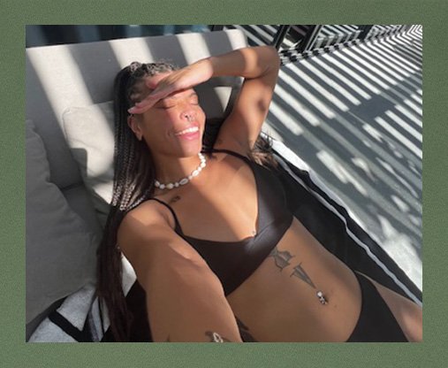 Aaliyah Ramsey lying down wearing a black bikini and taking a selfie in the sunlight, sheilding her eyes from the sun and smiling