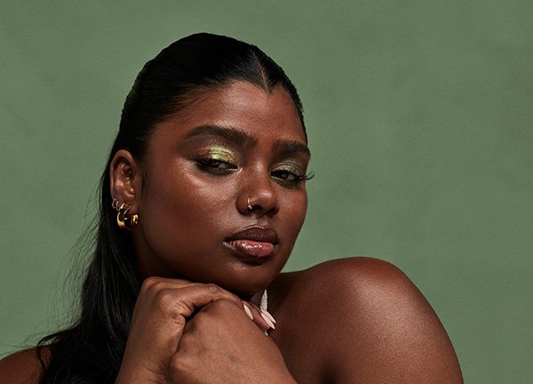 A close up of deep-toned model wearing green eyeshadow and lip gloss in front of a green background in a studio setting.