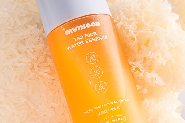 A close up shot of MUIHOOD’S bestselling Tao Rice Water Essence laying angled on a bed of rice and reflecting a orange-toned hue.