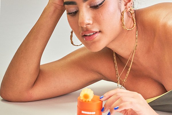 A wide shot of a female model playing with a Muihood Good Chi Cleansing Balm as she has her hand on her head in a studio setting.