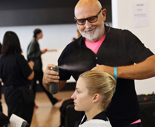 Sam McKnight is wearing a black rolled up jumper, holding a bottle of hairspray and spritzing a model’s slicked back hairstyle.