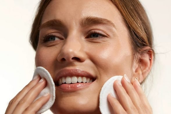 A close up of a female applying Caudalie’s face toner to her face with two cotton pads, in a bathroom setting, against a white background.