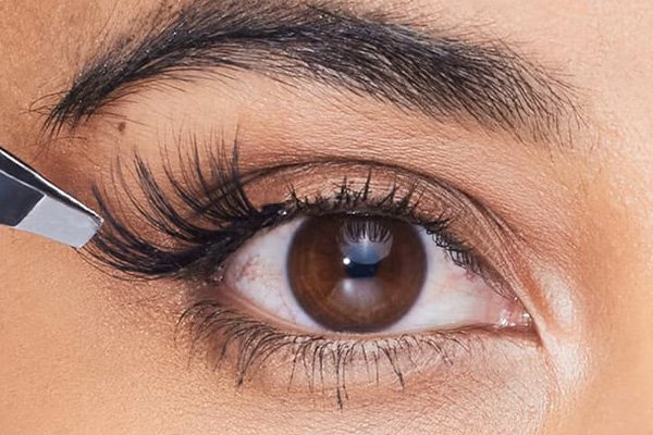 close up of a brown eyed model applying fake eyelashes to the outer corner of her eyes using a tweezer