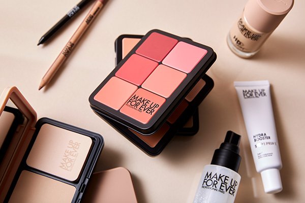 a collection of MAKE UP FOR EVER products including a stack of all in one palettes, lip lines, powder foundation and setting sprays. shot in a studio against a beige background