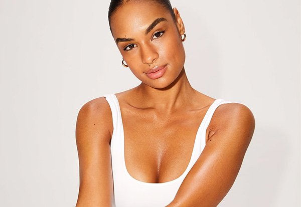 A model with tan skin, groomed brows and a pink lip wearing a white vest top and gold hoop earrings with her hair in a bun.