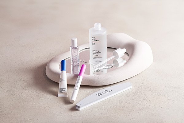 A flat lay image of Dr. Dana’s Nail Renewal System products sitting on a mirror, in a studio setting on a beige background.