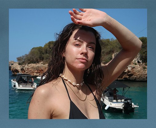 Maddie Edwards standing on a rock with the sea behing her. She is wearing a black bikini top and a shell necklace, and has her arm up to sheilf her eyes form the sun.