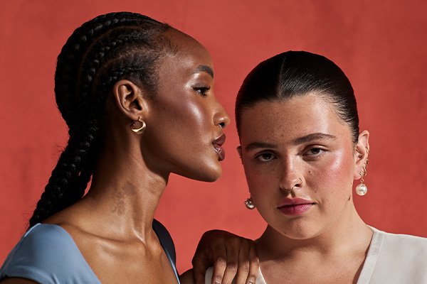 two models - one dark skinned, one pale with dark hair scraped back. Both models have glowy skin and the dark skinned model is posed sideways looking at the other model she is taller and wearing a blue tshirt. This image in shot against a dark coral background