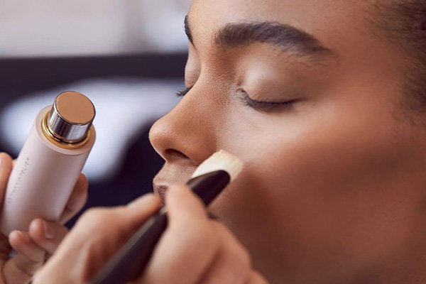 westman atelier vital skin foundation stick being allied on a dark skinned mode. the MUA is holding a foundation brush in one hand and the foundation stick in the other. the mode has thick eyebrows and her eyes are closed