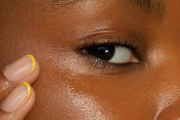 a close up of the eyes of a dark skinned model applying eyecream to her under eyes. her nails are short and she has thin french yellow tips on her nails.