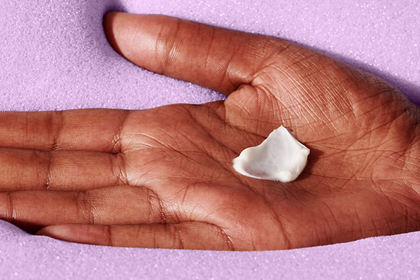 A medium shot of Drunk Elephant’s Lala Retro Whipped Cream in the palm of a model’s hand. In a studio setting, in front of a purple background. 