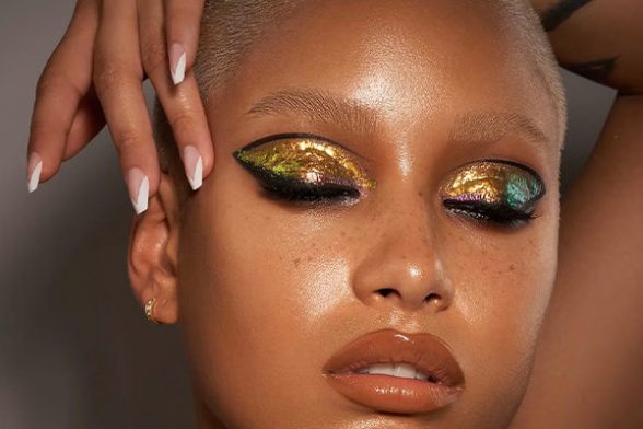 A close up of a model wearing gold and green glitter eyeshadow, black liquid eyeliner, with a brown lip and a French manicure