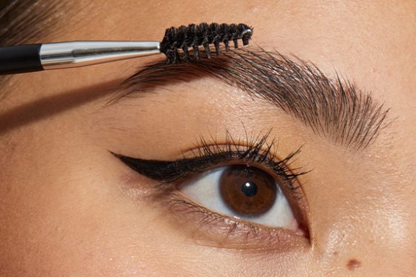 model with dark brown eyes and eyebrows using a spoolie to brush up her eyebrows