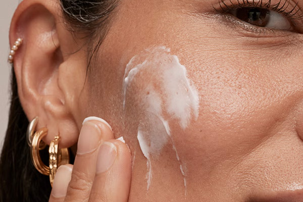 A close up of a female model rubbing a face moisturiser into her face, in a studio setting, in front of a white background.