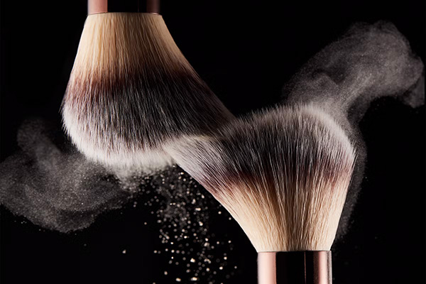 A close up image of two of Hourglass make up brushes hitting each other with pigment flying off it, in front of a black background.