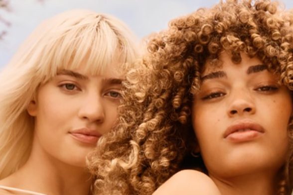 A close up shot of two female models. One with straight, blonde hair with a fringe and the other with brunette, curly hair with a fringe.