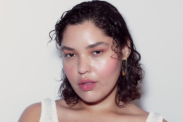 A medium shot of a female model with glossy, curly, short hair looking directly into the camera, wearing a white tank top. 