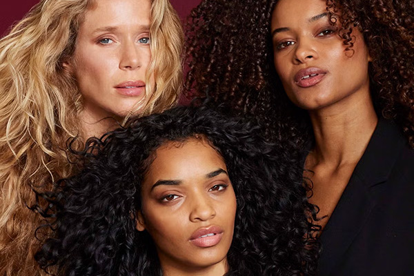 An image of 3 female models with different textured and coloured curly hair, in a black suit in a studio setting. Infront of a red background. 