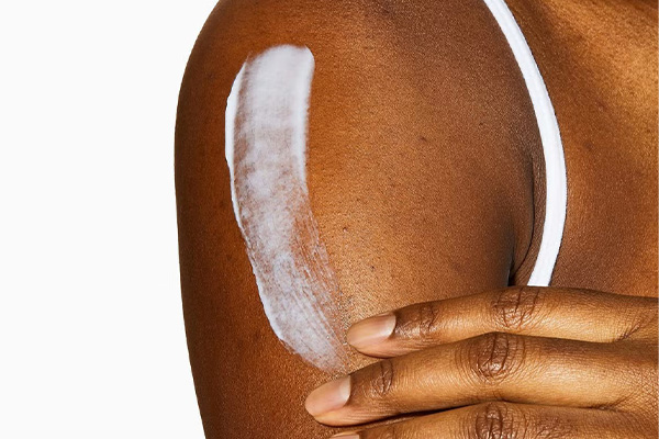 a dark skinned model applying a white cream over her shoulders. you can't see her face just arms and hands. shot in a studio