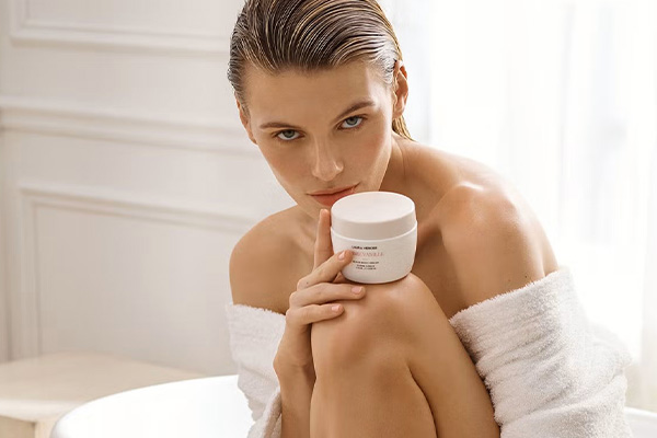 model in bathroom wearing a bathrobe with her shoulders exposed holding a tub of laura mercier body crea