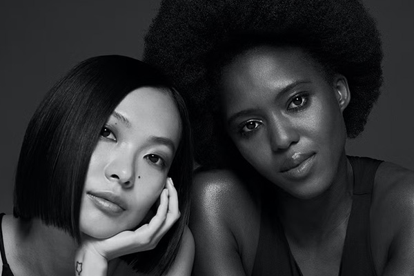 two models- one asian and one dark skinned are looking into the camera, shot in a studio. the asian model has a straight bob while the dark skinned model has an afro