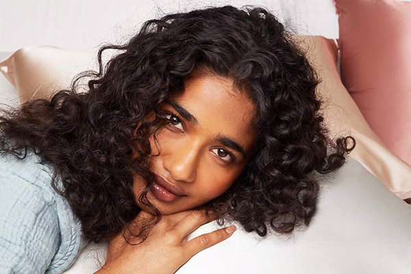 A close up of a female model with black curly hair laying her head down on a pile white and pink of silk pillows in bed.