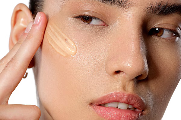 A close up shot of a female model applying a glutathione enriched serum to her cheek, in a studio setting, in front of a white background. 