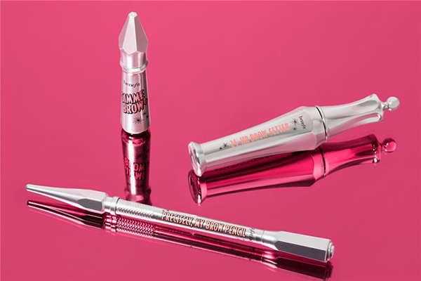 A wide shot of three Benefit eyebrow products laying randomly on a pink background, in a studio setting showing their reflections. 